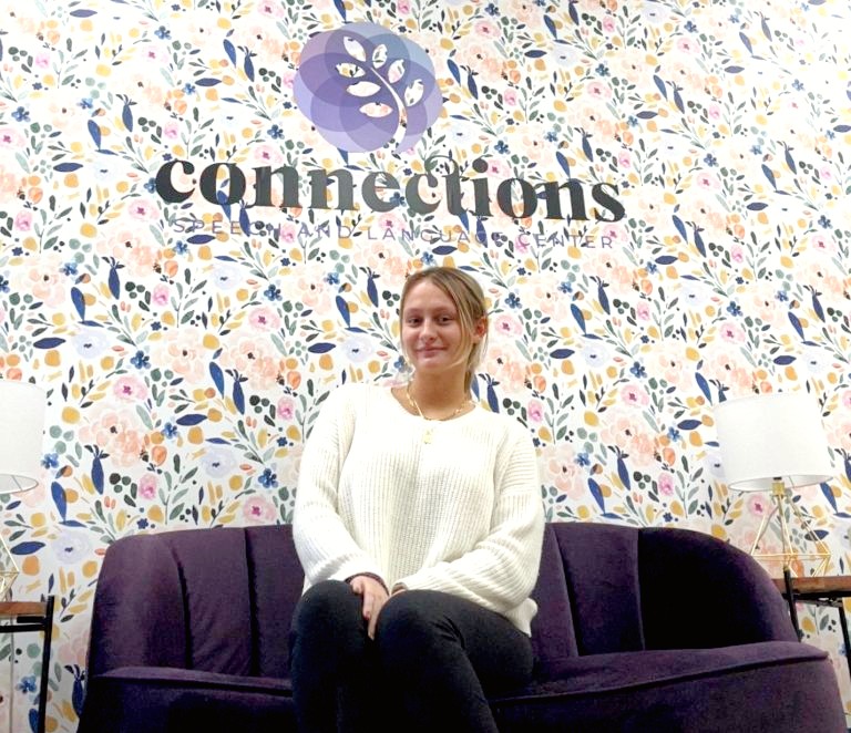 woman sitting on a purple couch wearing a white sweater. In the background is floral wallpater and the word "connections."