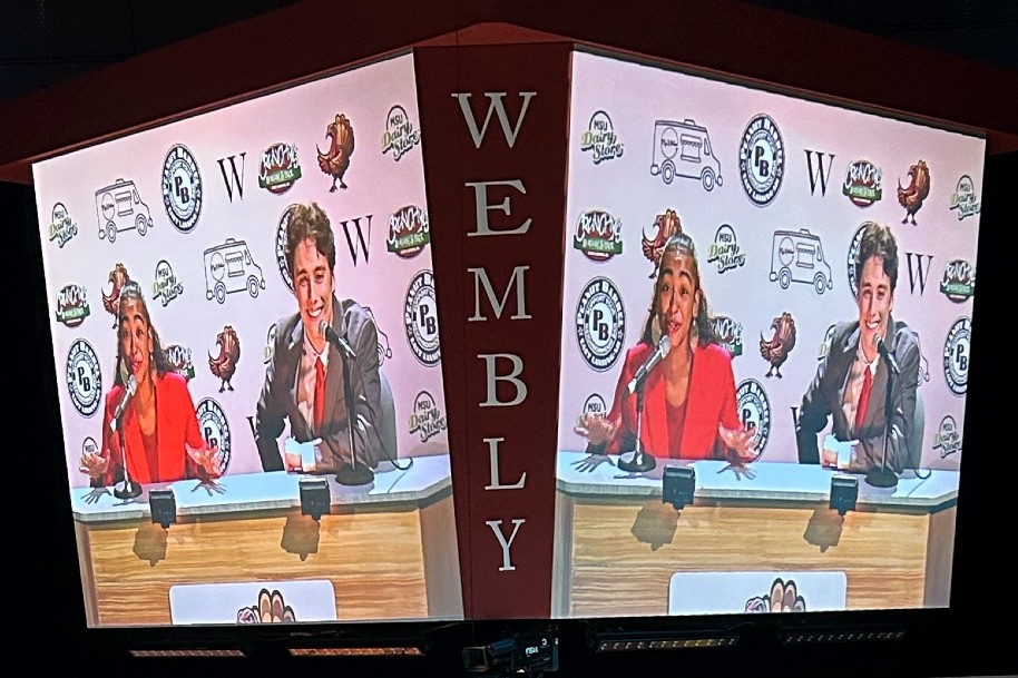 A photo showing two TV monitors and on each monitor the same image is appears of two sportscasters, one female on the left wearing a red suit and one man on the right wearing a gray suit with a red tie, both sitting at a desk talking into microphones. 