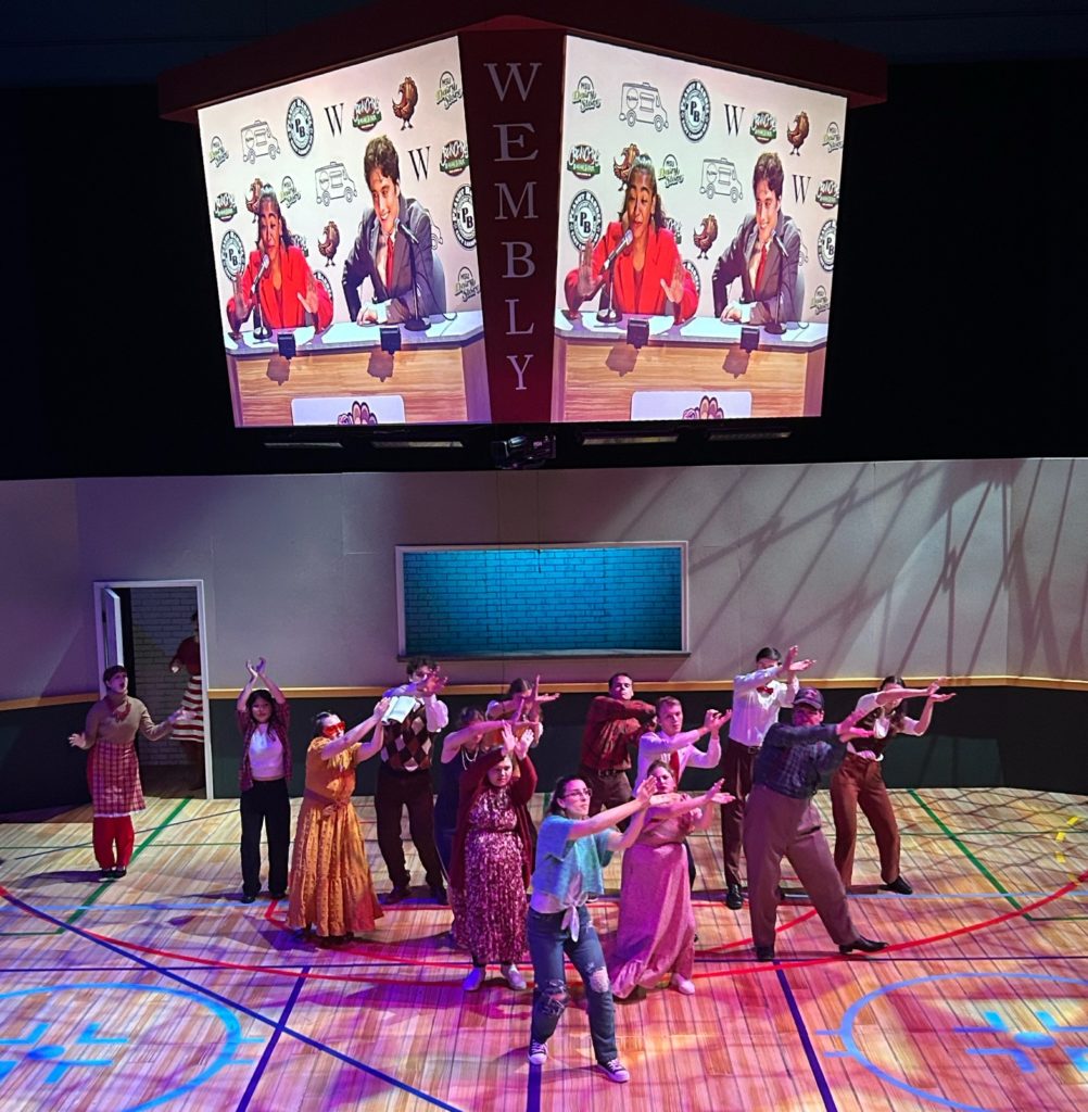 Photo of a group of 14 people on a stage doing a dance with a couple TV monitors overhead that both show two sports broadcasters. 