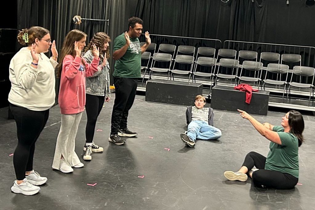 Four people standing on the left of the photo with both their hands raised. In the middle is a man leaning back on the ground with one of his legs crossed and on the right of the photo is a woman itting on the ground with her legs crossed staring and pointing at the four people who are standing. 