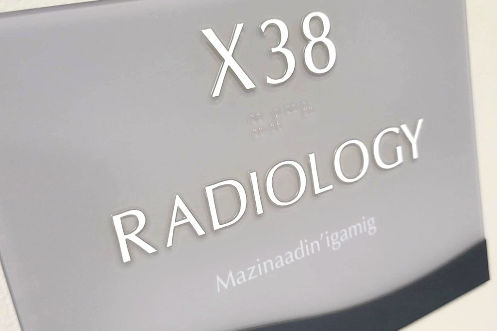 A photo of a room number nameplate at a hispital. The nameplate says: (X38 Radiology Mazinaadin'igamig. 