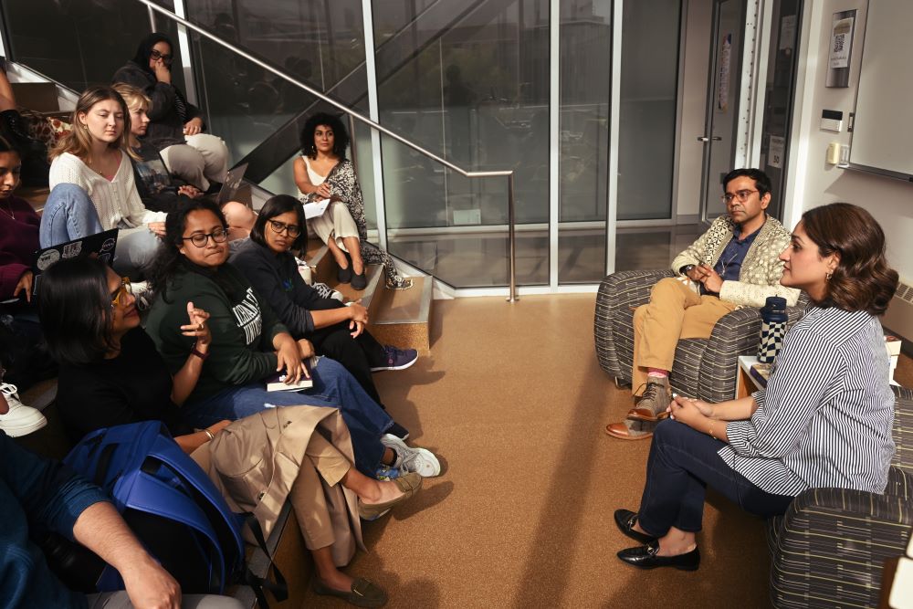 Two people sit at the front of a room in an auditorium setting. A person in the front row of the crowd asks a question.