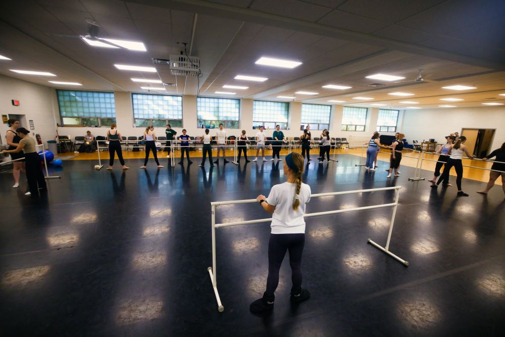 A dance studio is filled with students at ballet bars as they watch the dance instructor in the front of the room.