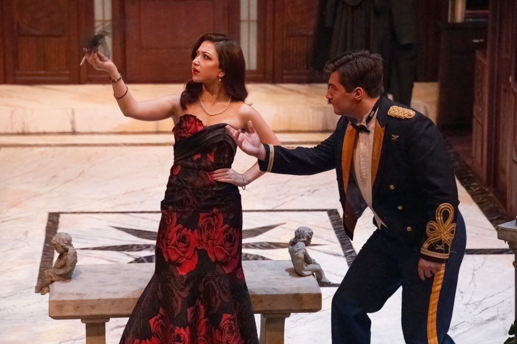 A woman wearing a long black sleeveless dress with red roses print and pearl knecklace who is looking at her raised hand that is holding something. A man wearing a military suit is standing next to her and is also looking at her raised hand. 