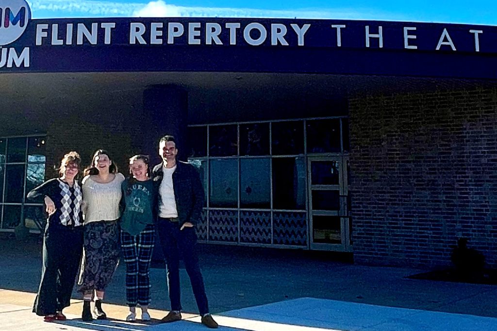 Four people standing together outdoors in front of the Flint Repertory Theatre building. A brick wall and windows can be seen in the background. The group poses casually and smiles.