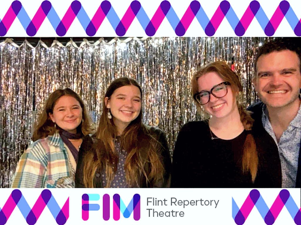 A picture of four individuals standing in front of a silver tinsel backdrop. They are smiling. The Flint Repertory Theatre branding and graphic frames the photo at the top and bottom.