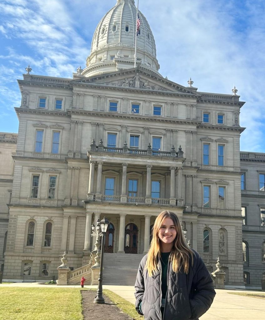 Woman with long blonde hair and wearing a black coat standing in front of the Michigan State Capitol building. 