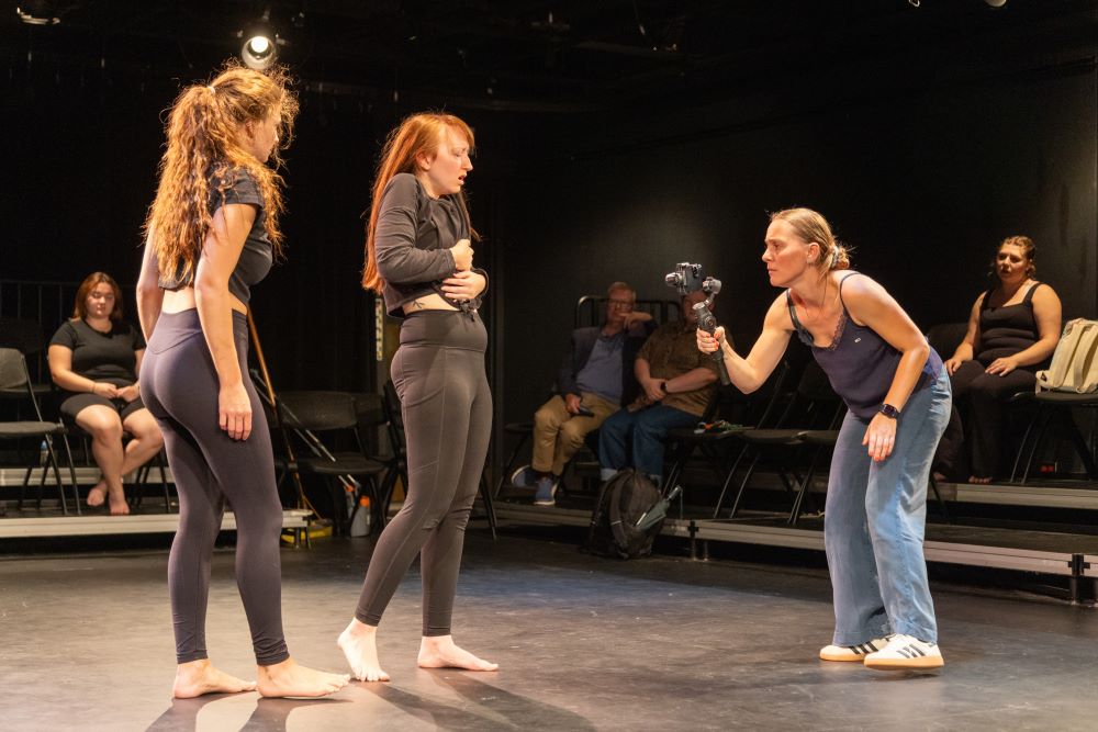 A person with a phone films two students while they rehearse dance movements on a black theatre stage.