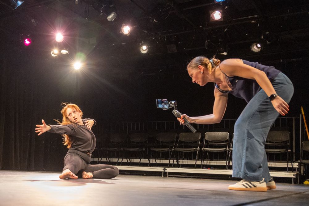 A person with a phone films a student as she hits her final dance move on the floor of a black theatre stage.