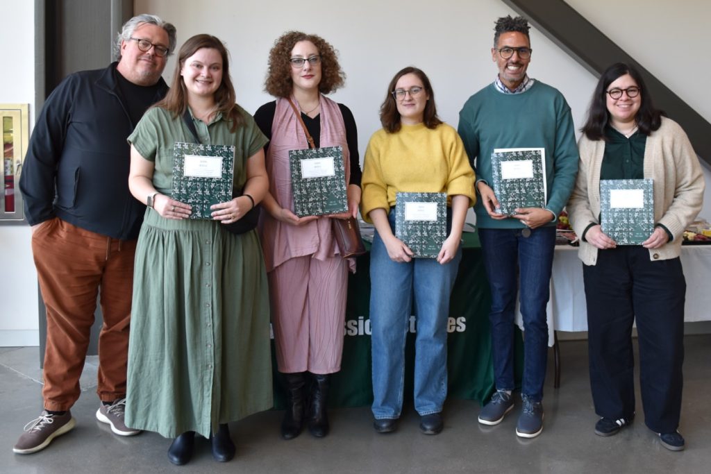 Six people standing in a row with the five people o the right holding the TROPOS journal. The people from left to right are: a man wearing glasses and a black shirt, a woman in a green dress, a woman wearing glasses and a rose-colored outfit, a woman wearing glasses a yellow shirt and jeans, a man wearing a green shirt and glasses, and a woman wearing glasses and a beige cardigan.