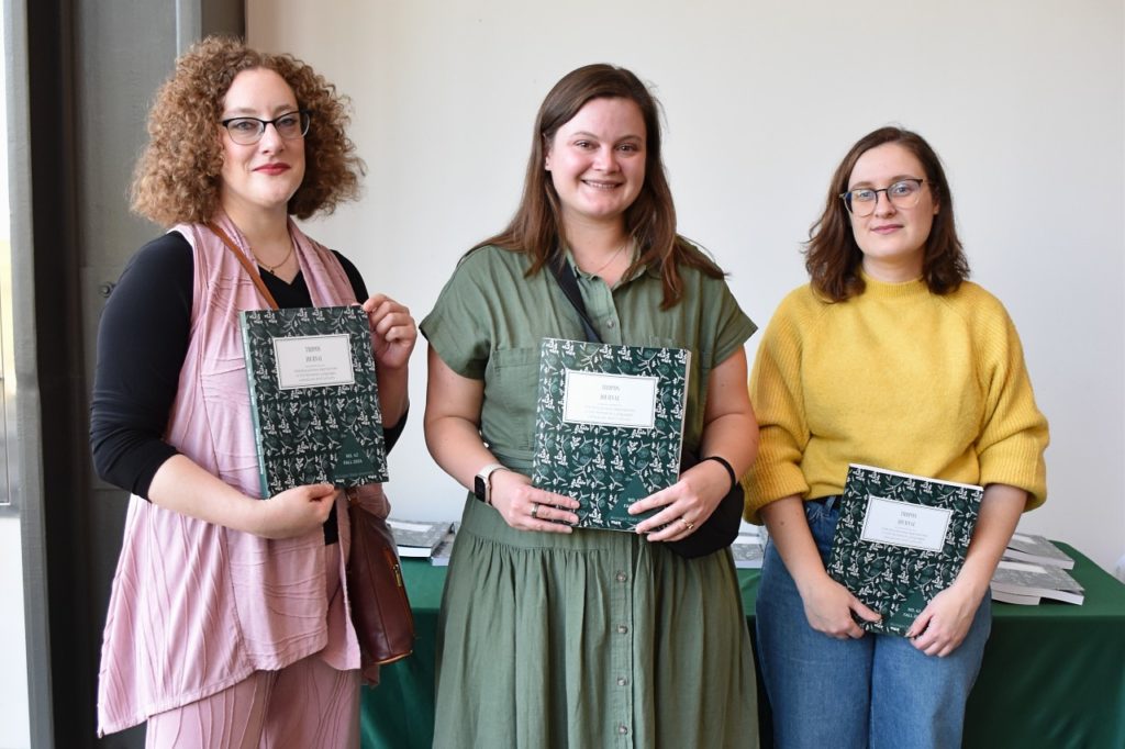 Three women standing in a row next to each other, each of them holding a copy of the TROPOS journal. The woman on the left has shoulder-length curly hair and is wearing glasses and a rose-colored outfit. The woman in the center is wearing a light green dress. The woman on the right is wearing glasses a yellow shirt and blue jeans. 