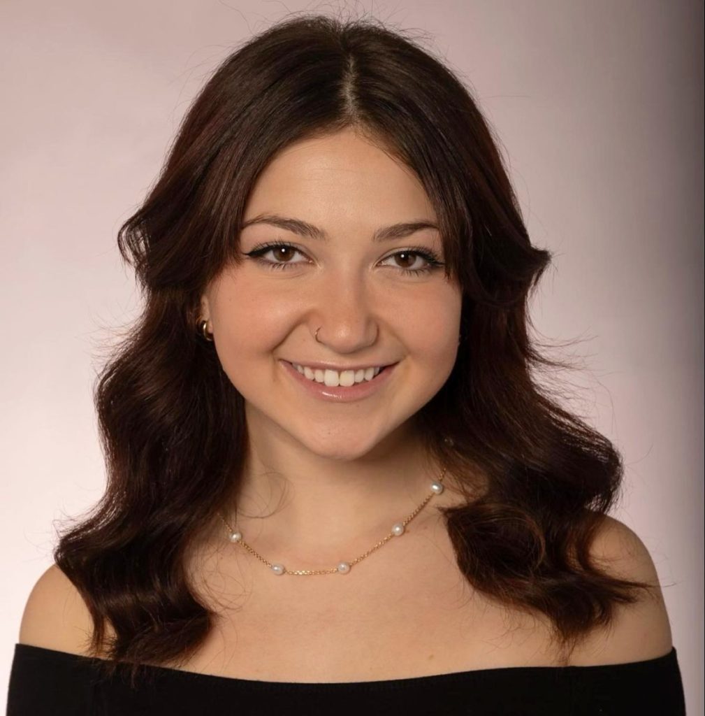 headshot of a woman who is smiling with long brown hair and a knecklace with pearls. 