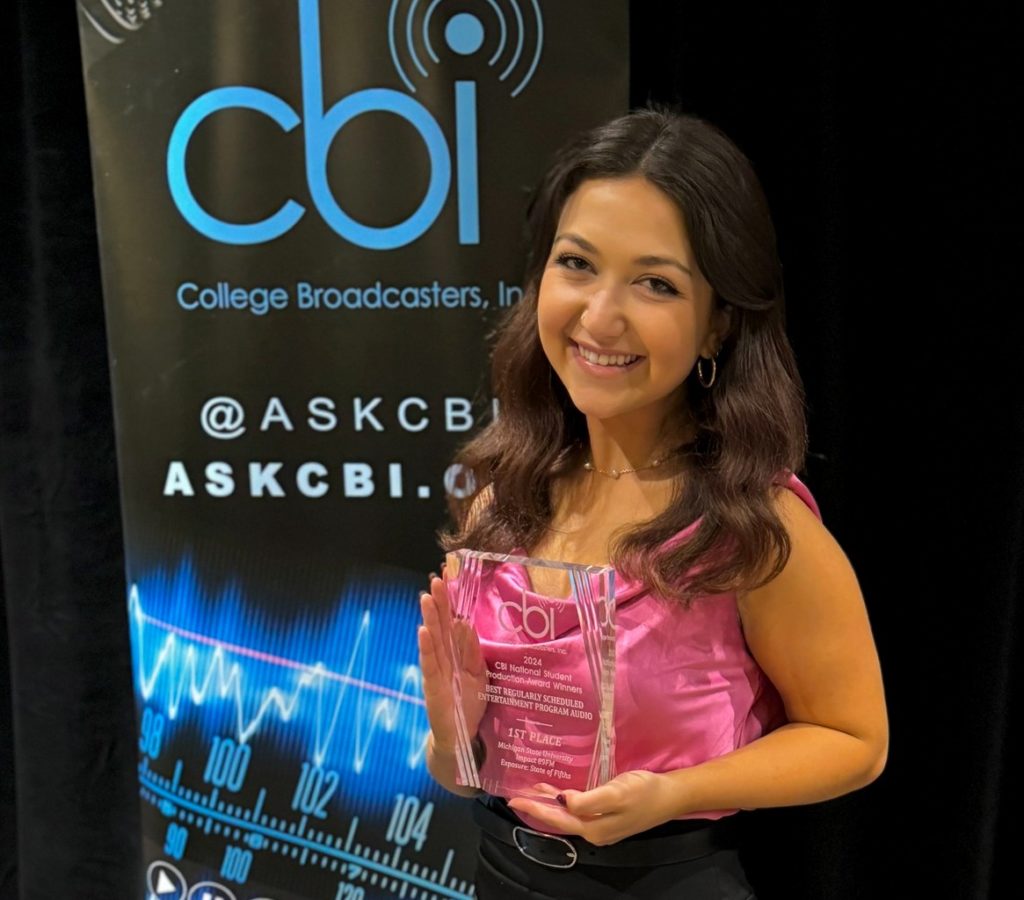 A woman with long brown hair and wearing a pink tank top is holding a trophy and behind her is a banner that says: " College Broadcasters, Inc."