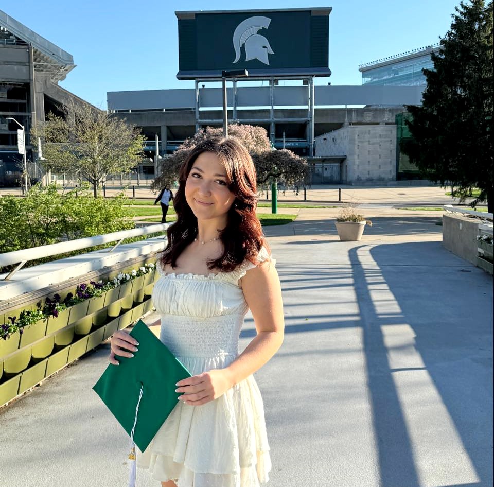 A woman with long brown hair and holding a green graduation cap, standing on the bridge next to Spartan stadium with the large Spartan helmet logo at the stadium in the background.