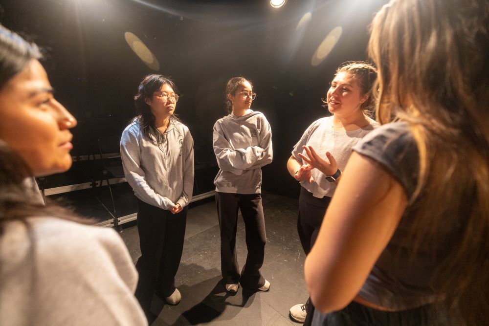 An angle in the midst of five students talking on a black theatre stage.