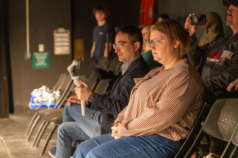 Two people watch a performance in a dark theatre setting. One is also filming the performance.