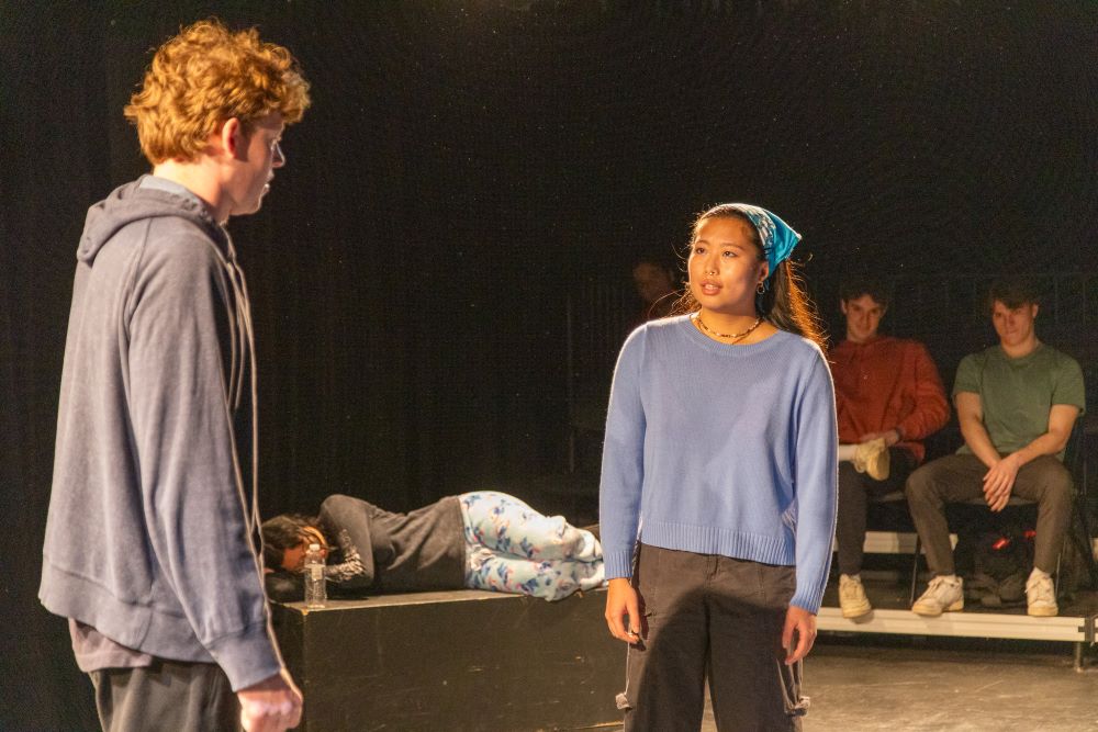 Two people give a monologue on stage in a dark theatre setting.