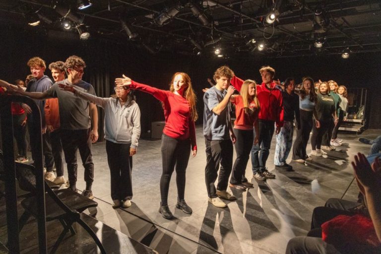 A group of performers are lined up in a V-shape on a black theatre stage.