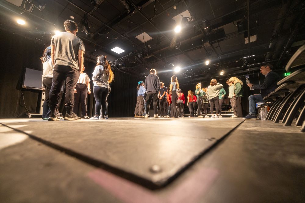 A low angle looking up at a group of students standing on a black theatre stage.