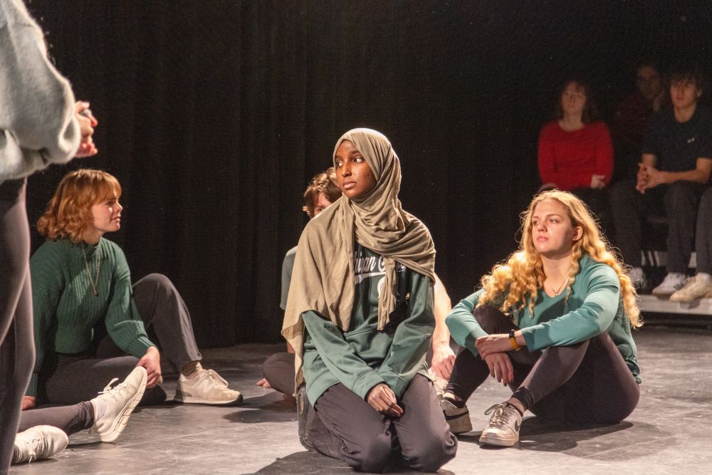 A group of students sit on the floor and listen as part of a performance in a dark theatre setting.