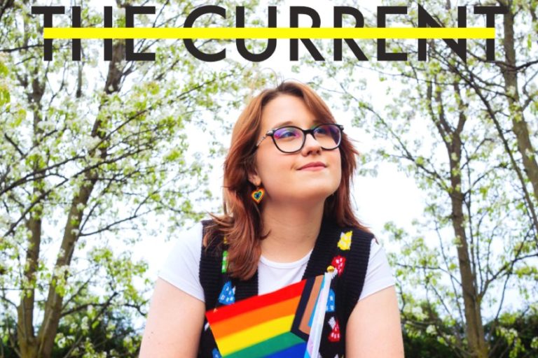 A picture of a magazine cover, with "The Current" written in bold black text with a yellow bar through the middle of the letters. A person with shoulder-length auburn hair looks into the distance, and holds a Pride flag.