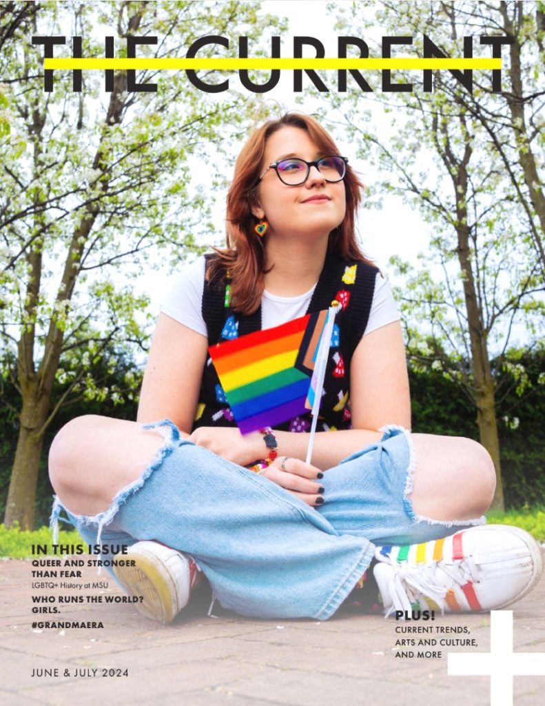 A picture of a magazine cover, with "The Current" written in bold black text with a yellow bar through the middle of the letters. A person with shoulder-length auburn hair looks into the distance, and holds a Pride flag. 