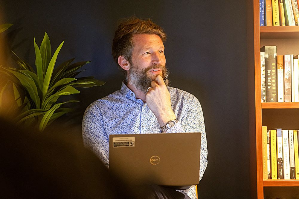 A man with a laptop sitting next to a bookcase looks to the left.