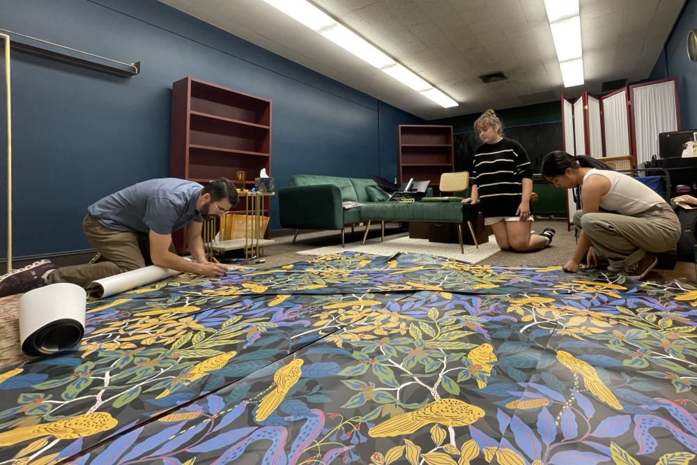 Three people lay out wallpaper on the floor to prepare it to paste on a wall. The wallpaper has a tree and bird design.