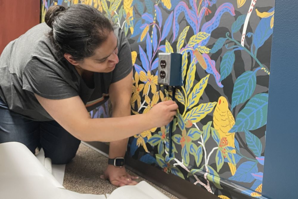 A person smooths wallpaper around a wall outlet. The wallpaper has a tee and bird design.