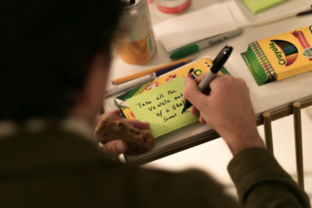 A person holds a cookie in one hand while in the other hand the person uses a marker to write on a Post-It note the writing prompt: "Take all the vowels out of a Shakespeare sonnet."