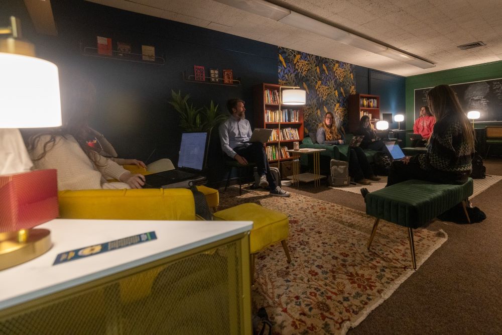 A professor with a group of students with laptops sit in cozy chairs in the MSU Writers' Studio space.