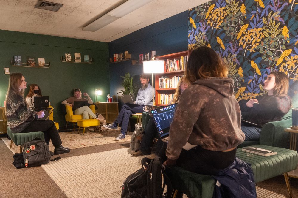 A professor with a group of students with laptops sit in cozy chairs in the MSU Writers' Studio space.