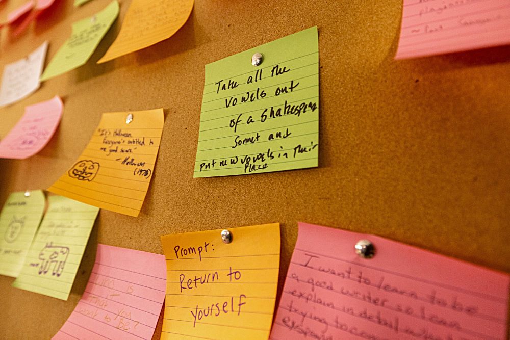 Writing prompts on Post-It notes are thumbtacked to a cork board.