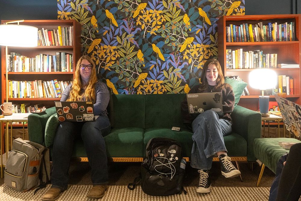 Two students with laptops sit on a cozy green couch in the MSU Writers' Studio on campus. In the background is a wallpapered wall showing trees and birds and two bookcases with books.