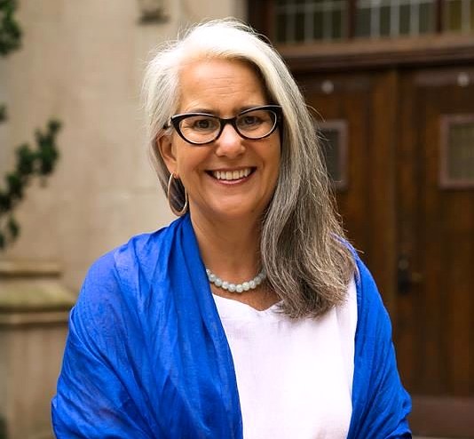 A woman with long gray hair, glasses, and a bright smile stands outside a building with wooden doors. She is wearing a blue shawl, a white blouse, and a pearl necklace.