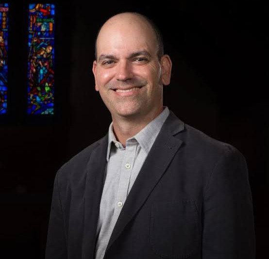 A man with a shaved head and a light gray shirt smiles. He is wearing a dark blazer, standing in front of a stained-glass window.