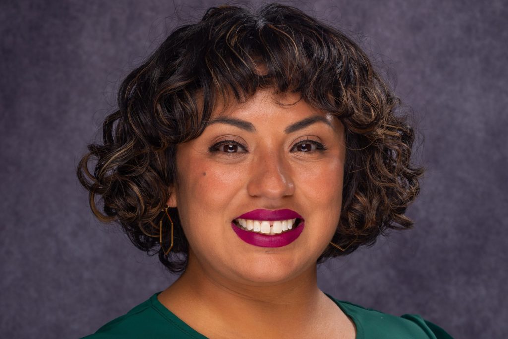 A close-up portrait of a person with short, wavy dark hair, wearing a green blouse and bright magenta lipstick, smiling in front of a neutral gray background.