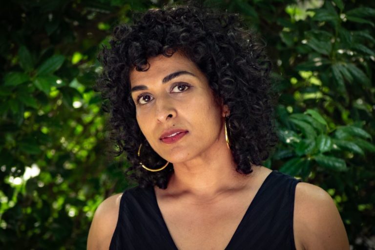 A person with curly hair, gold hoop earrings, and a black shirt stands outside in front of green foliage.