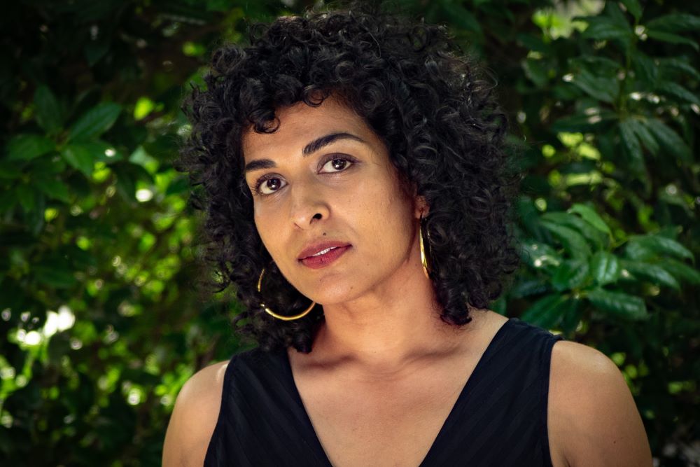 A person with curly hair, gold hoop earrings, and a black shirt stands outside in front of green foliage.