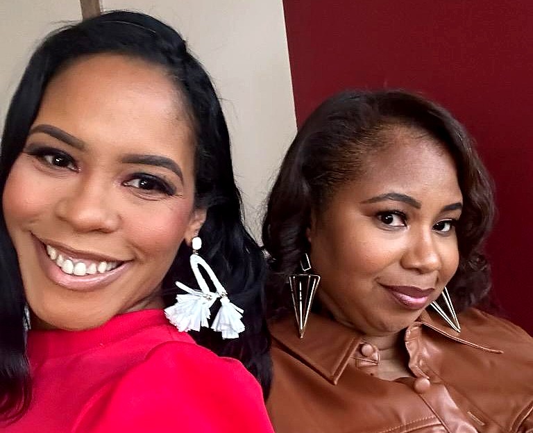 Two Black women standing next to each other. Both are wearing large earrings with the woman on the left wearing a bright pink shirt and the woman on the right wearing a brown leather shirt. 