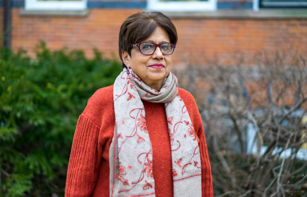 A picture of a woman with short, brown hair, and pink glasses. She wears a burnt orange sweat and grey scarf embroidered with red floral patters. In the background, there is shrubbery and a brick wall. 