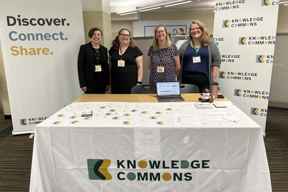 Four people stand behind a table that has brochures and a laptop on it. There’s a white tablecloth that says: “Knowledge Commons.” Behind the people are two large banners. The banner on the left says: “Discover. Connect. Share.” The banner on the right says: “Knowledge Commons” repeated across the banner.