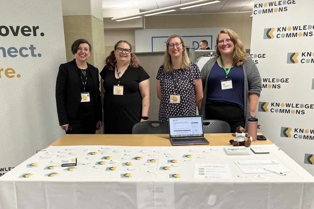 Four people stand behind a table that has brochures and a laptop on it. There’s a white tablecloth that says: “Knowledge Commons.” Behind the people are two large banners. The banner on the left says: “Discover. Connect. Share.” The banner on the right says: “Knowledge Commons” repeated across the banner.