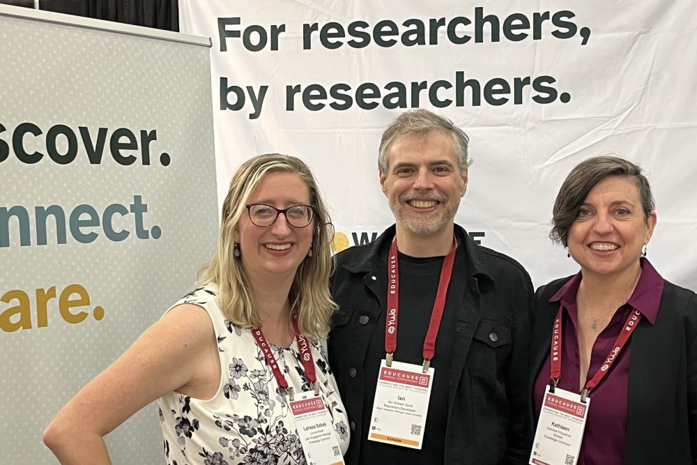 Three people stand in front of two banners. The banner on the left says: “Discover. Connect. Share.” The banner behind the people says: “For researchers, by researchers.”