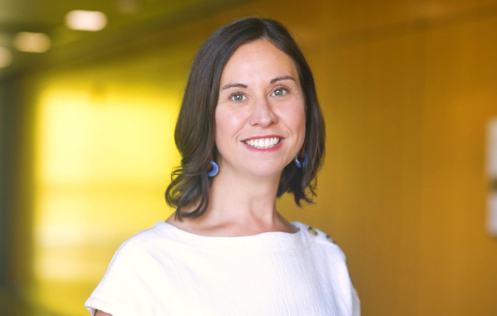 A woman with shoulder-length dark hair and blue earrings smiles brightly. She is standing in a brightly lit hallway with a yellow background, wearing a white blouse.