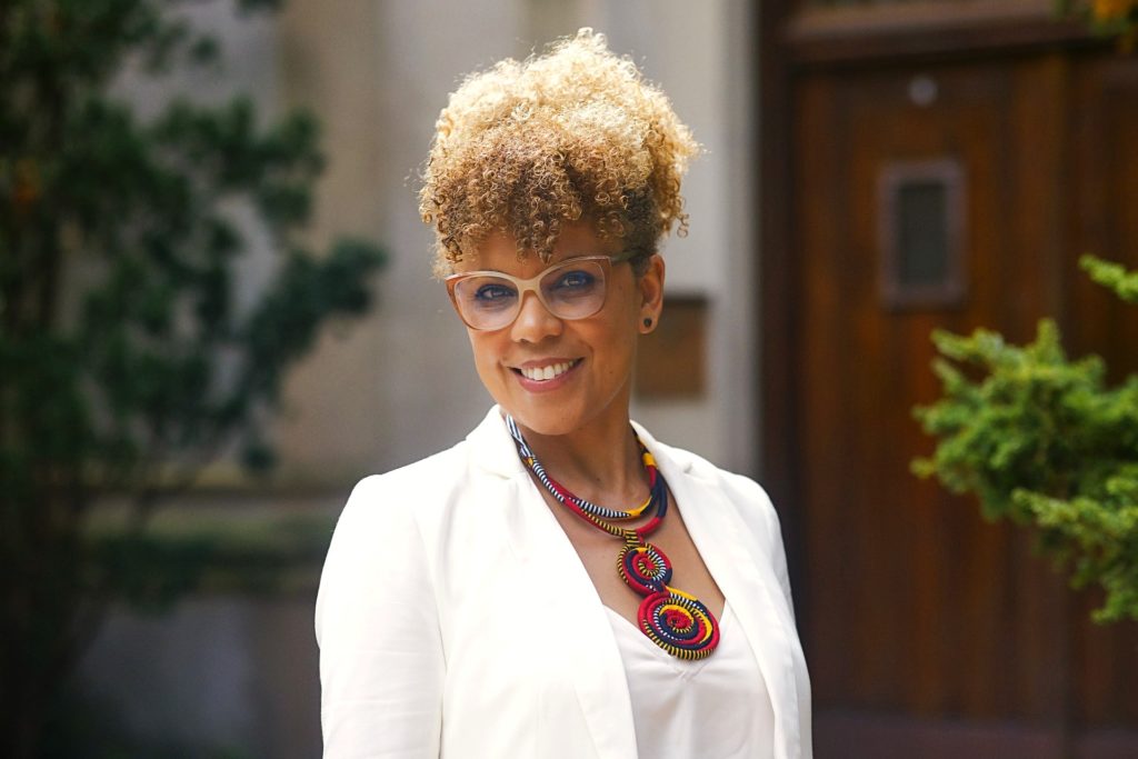 A picture of a person with curly brown and blond hair styled in an updo, wearing glasses, a white blazer, and a colorful, multicolored beaded necklace. The background includes greenery and a wooden door.