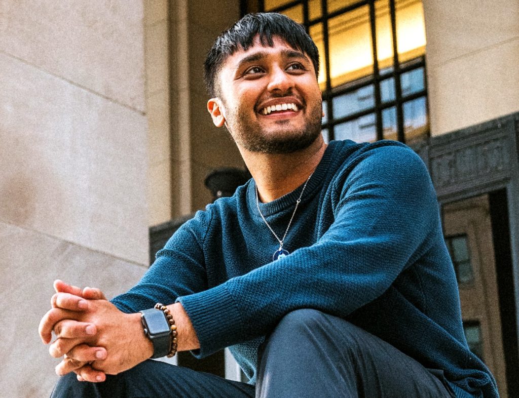 Man with short black hair and a short beard smiling and sitting with his arms resting on his knees and his hands clasped together. He is wearing a dark teale blue sweater and gray pants. 