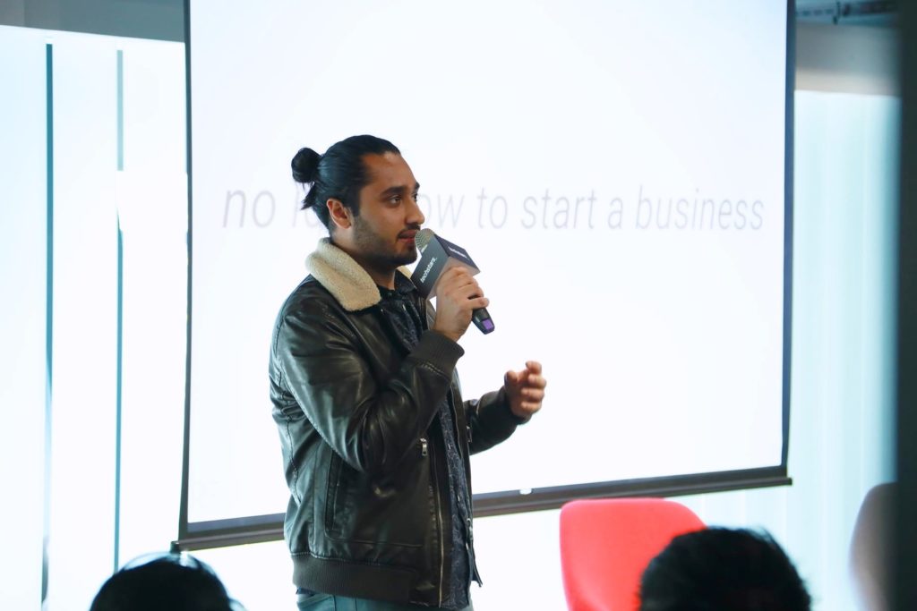 A picture of a speaker holding a microphone presenting in front of a projector screen. The individual wears a leather jacket with a shearling collar and has their hair tied back. An audience is visible in the foreground.