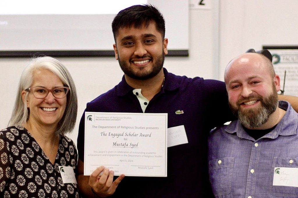 Three people standing in a row. The person on the left is a woman with shoulder-length gray hair and glasses. The person in the center is a man with dark black hair and a beard and moustache who is holding a certaificate that says: "The Engaged Scholar Award to Mustafa Syed", and the person on the right is a man who is bald and has a beard and moustache. 