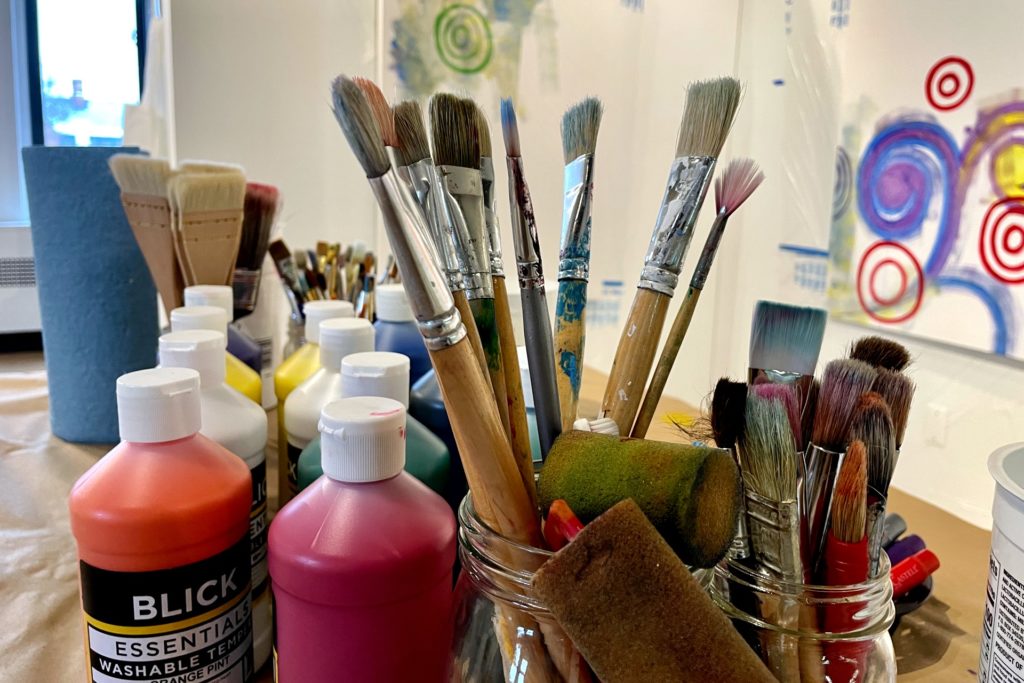  A close-up photo of an art station with jars of paintbrushes and sponges, surrounded by bottles of paint. Abstract artwork on cream walls is visible in the background